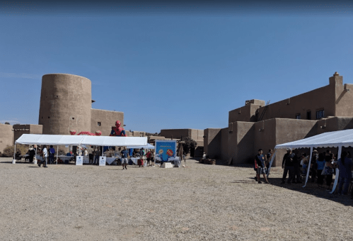 A desert scene with tents set up for an event near adobe-style buildings and a round tower under a clear blue sky.