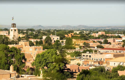 A panoramic view of a city with adobe-style buildings, greenery, and distant mountains under a clear blue sky.