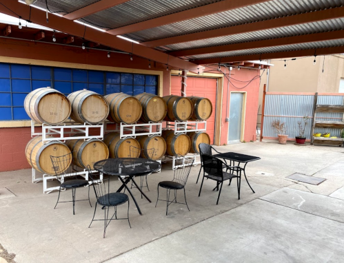 A rustic outdoor space with wine barrels stacked against a wall and a small table and chairs set up nearby.