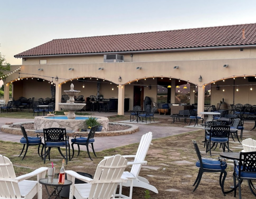 Outdoor seating area with white chairs, a fountain, and string lights, set against a building with a tiled roof.