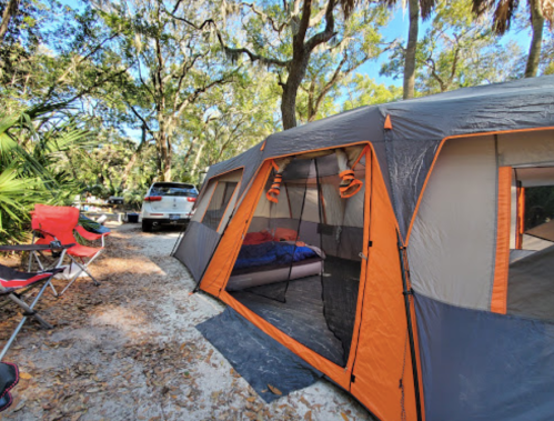 A spacious tent with orange accents set up in a wooded area, with camping chairs and a car nearby.
