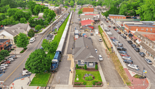 Aerial view of a train station with a train, surrounded by shops and greenery in a small town.