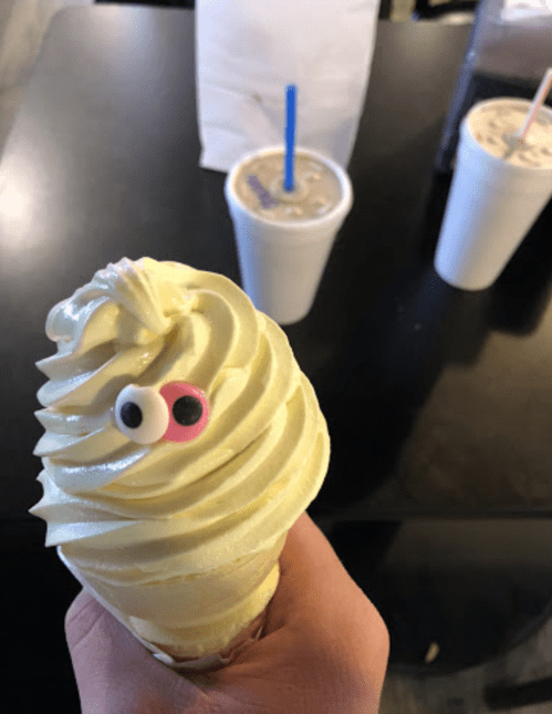 A hand holding a soft-serve ice cream cone with a playful googly eye on top, with drinks in the background.