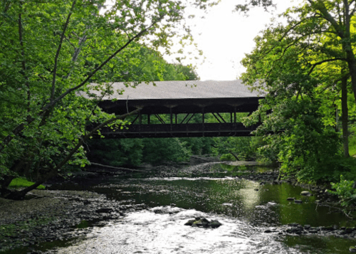 A covered bridge spans a serene river, surrounded by lush green trees and a peaceful landscape.