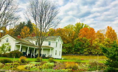 A white house surrounded by colorful autumn trees and a garden, under a cloudy sky.