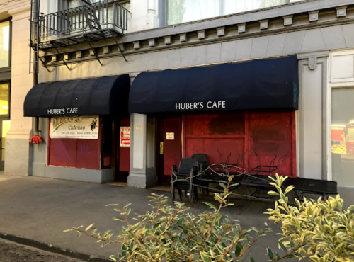 Exterior of Huber's Cafe featuring a dark awning and red-tinted windows, with a bench in front and plants nearby.
