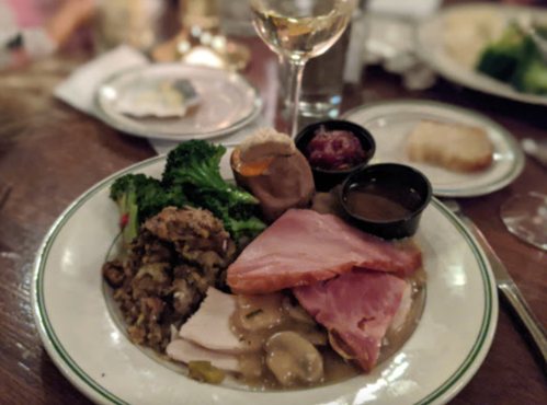 A plate of holiday dinner featuring turkey, ham, stuffing, broccoli, gravy, and sides, with a glass of white wine.