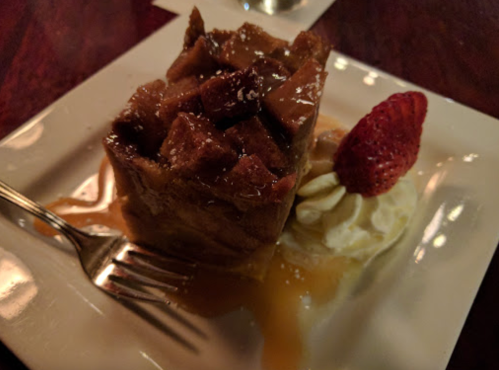 A slice of bread pudding topped with caramel sauce, a strawberry, and whipped cream on a white plate.