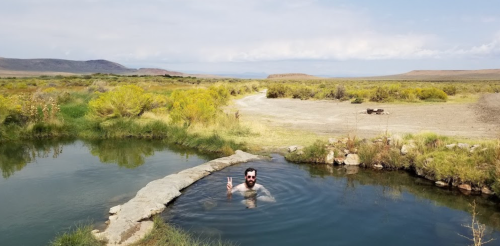 A person with a beard relaxes in a natural hot spring surrounded by a scenic landscape of grass and distant mountains.