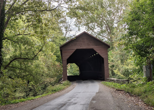 A wooden covered bridge surrounded by lush green trees, with a road leading into its entrance.