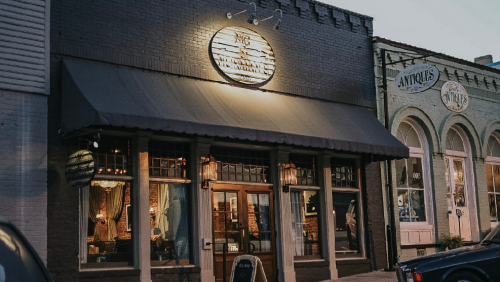 A charming storefront with a lit sign, featuring large windows and an awning, set in a quaint street.