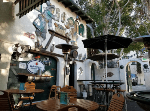 Outdoor seating area of a restaurant with murals, patio heaters, and umbrellas under trees.