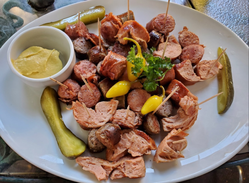 A platter of assorted meats, including sausages and pulled pork, garnished with pickles, mustard, and herbs.