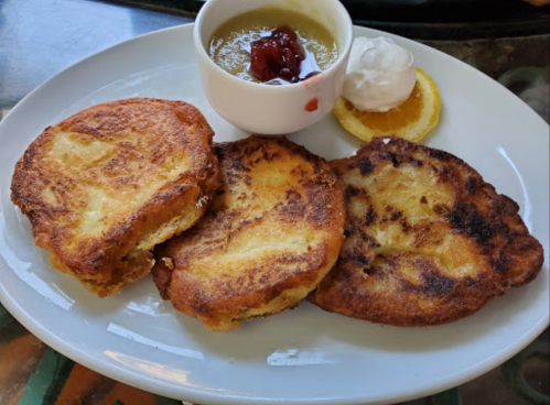 Three golden-brown fried pancakes on a plate, served with a small bowl of fruit compote, whipped cream, and an orange slice.