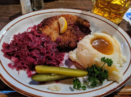 A plate with breaded meat, mashed potatoes with gravy, red cabbage, pickles, and a lemon wedge, alongside a beer.