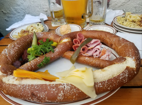 A large pretzel platter with assorted meats, pickles, cheese, and garnishes, served with beer in the background.