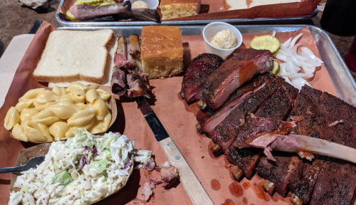 A platter of barbecue ribs, macaroni and cheese, coleslaw, cornbread, pickles, onions, and slices of bread on a tray.