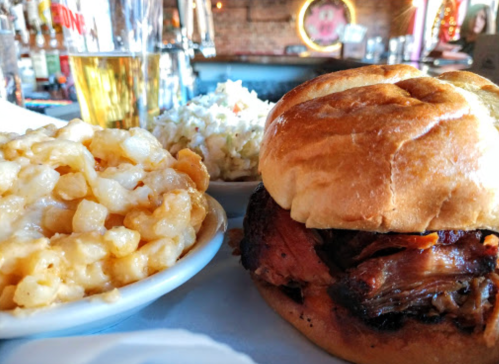 A plate featuring a pulled pork sandwich, macaroni and cheese, and coleslaw, with a beer in the background.