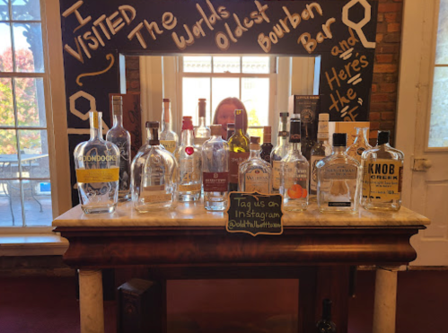 A display of various bourbon bottles on a bar, with a sign reading "I visited the world's oldest bourbon bar."