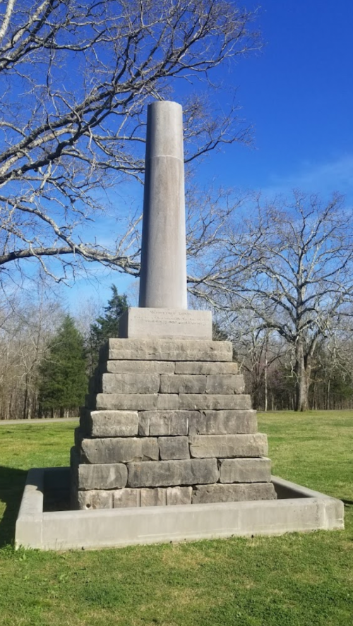 A tall stone monument on a stepped base, surrounded by grass and bare trees under a clear blue sky.