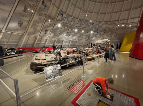 A museum exhibit featuring a damaged car, with visitors exploring the space and historical portraits on the walls.