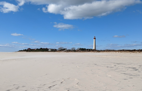 A tall lighthouse stands on a sandy beach under a blue sky with scattered clouds.
