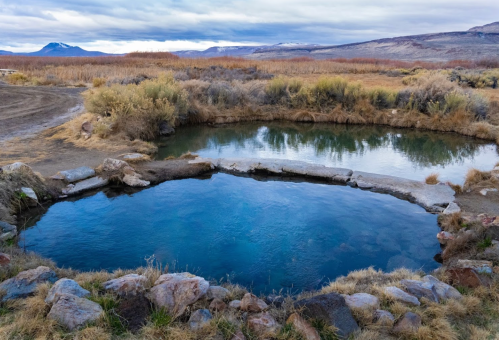 Two natural hot springs surrounded by rocky edges and sparse vegetation, set against a backdrop of mountains and cloudy skies.