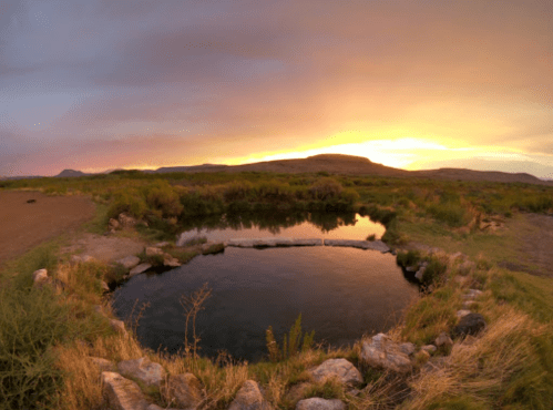A serene hot spring surrounded by grass and rocks, reflecting a colorful sunset in the background.