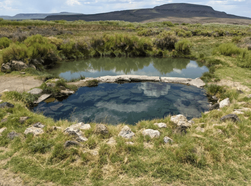 A natural hot spring surrounded by grass and rocks, with mountains in the background under a cloudy sky.