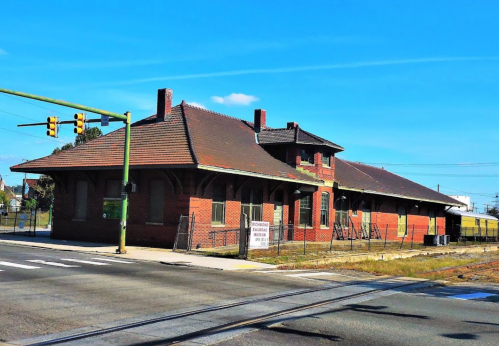 Historic brick train station with a tiled roof, surrounded by a fence and located at a street intersection.