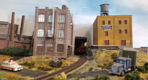 A miniature train scene featuring buildings, a train entering a tunnel, and toy vehicles on a scenic layout.