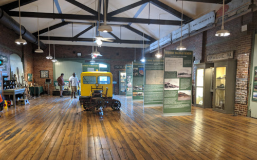 Interior of a museum with wooden floors, exhibits, and a vintage yellow vehicle on display. Two visitors are exploring.