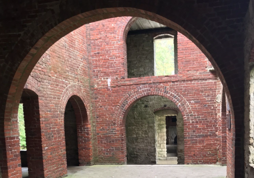 Interior of a brick ruin featuring arched doorways and windows, with stone walls and a natural light source.