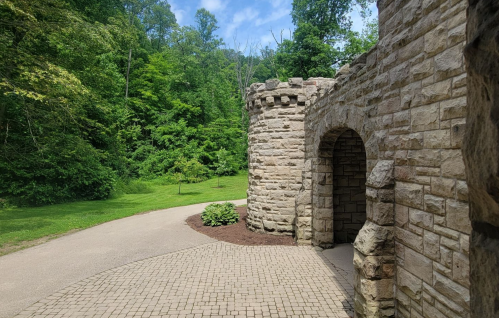 A stone castle turret beside a paved path, surrounded by lush green trees and a clear blue sky.