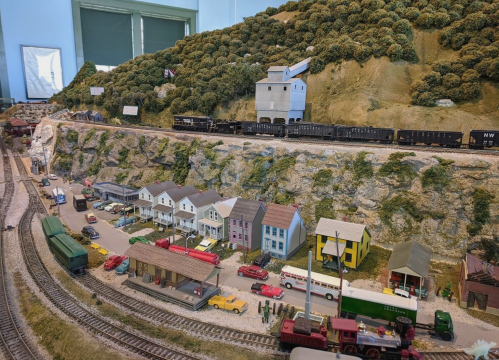 A detailed model train layout featuring a hillside, buildings, and miniature vehicles along the tracks.