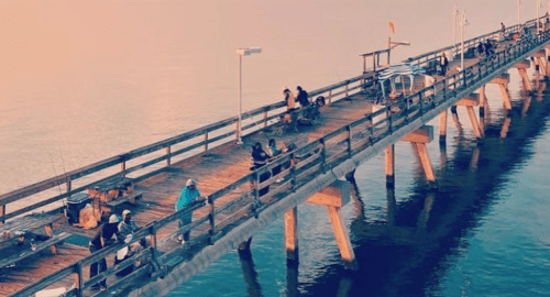 A wooden pier extends over calm water, with people fishing and enjoying the view at sunset.
