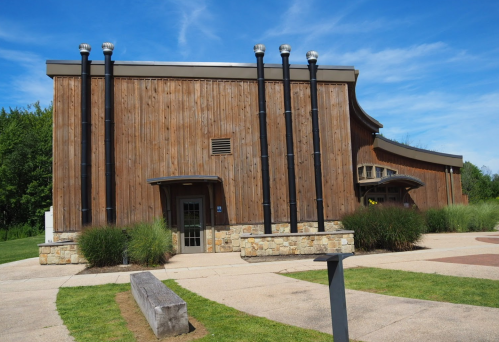 A modern wooden building with stone accents, surrounded by greenery and a clear blue sky.