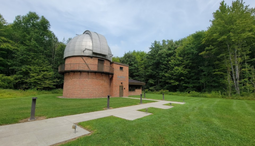 A brick observatory with a dome, surrounded by green trees and a grassy area, under a cloudy sky.