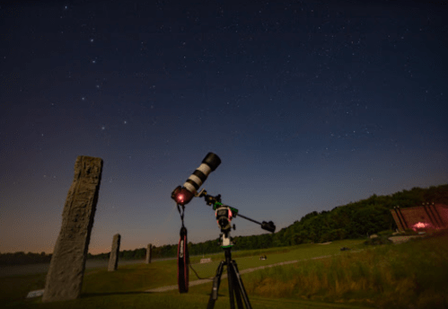 A telescope on a tripod points towards a starry sky, with stone monoliths visible in the foreground.