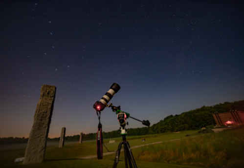 A telescope on a tripod points towards a starry sky, with stone monoliths visible in the foreground.