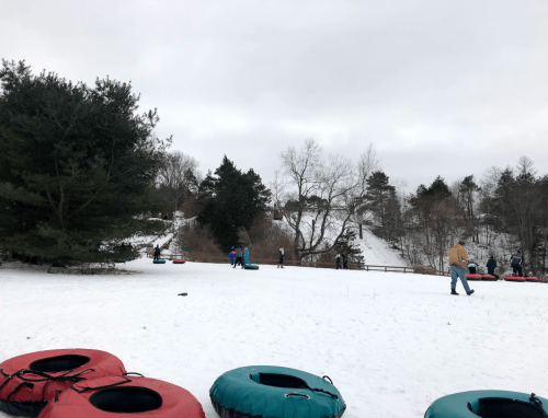 A snowy hill with people tubing and walking, surrounded by trees and a cloudy sky. Colorful tubes are in the foreground.