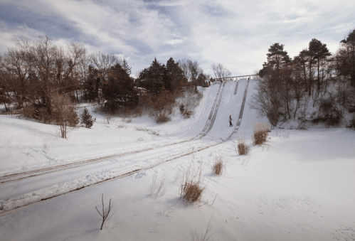 A snowy landscape with a sledding hill and a person sledding down, surrounded by trees and a cloudy sky.
