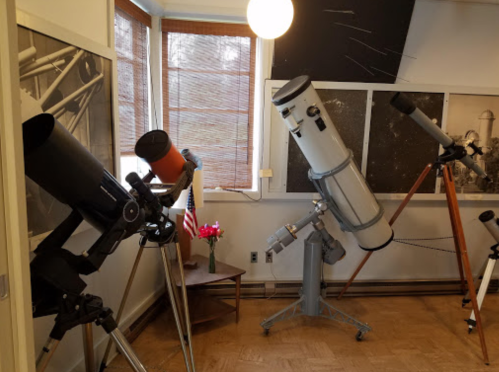 A room with several telescopes positioned towards a window, with a small table and flowers in the background.