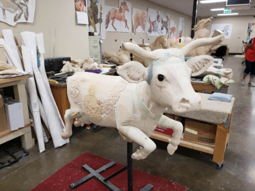 A sculpted cow model on a stand, surrounded by art supplies and animal sketches in a workshop setting.