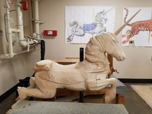 A partially carved wooden unicorn sculpture in a workshop, with sketches of a unicorn and a tiger on the wall.