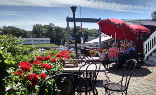 Outdoor dining area at a waterfront restaurant, featuring red flowers, umbrellas, and patrons enjoying the view.