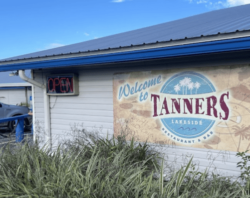 A sign for Tanners Lakeside Restaurant Bar, featuring "Welcome" and "Open" in bright neon. Lush greenery in the foreground.