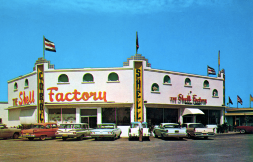 A vintage building with "The Shell Factory" sign, featuring classic cars parked outside and flags on display.