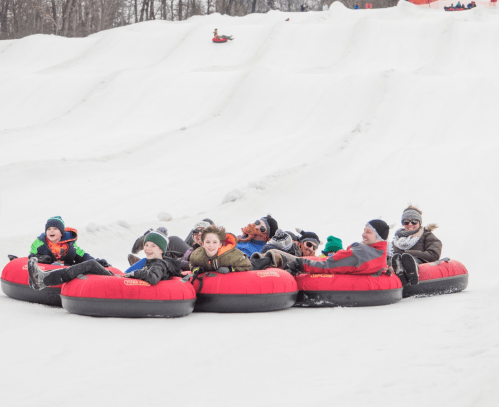 A group of children and adults enjoying snow tubing on a snowy hill, smiling and having fun in red inflatable tubes.