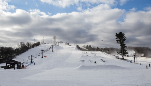 A snowy ski hill with a ski lift, surrounded by trees and cloudy skies, with people skiing and snowboarding.